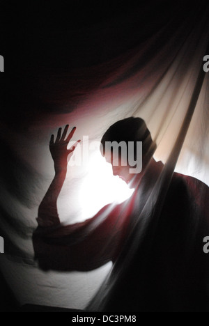 Silhouette of man behind curtain. The light picks up the red in his shirt and tinges the sheet with red light. Stock Photo