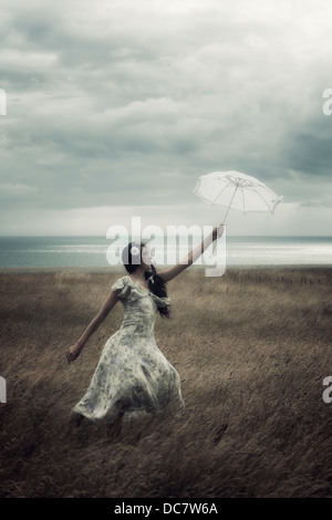 a girl in a floral dress on a field with a parasol is fighting against the wind Stock Photo