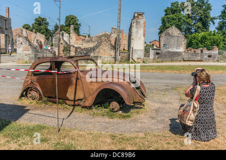 A woman taking a photograph of the iconic, left-to-rust 'Doctor's Car' among the ruins of Oradour-sur-Glane, war massacre village, Limousin, France. Stock Photo