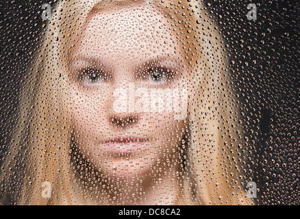 Portrait of young blond female teenager looking out of window covered with raindrops Stock Photo