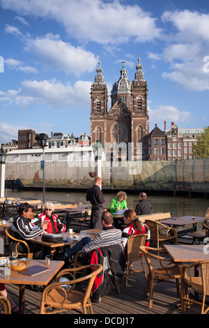 Tourists having lunch in cafe restaurant by the canal in Amsterdam, Netherlands, St Nicholas Church in the background. Stock Photo