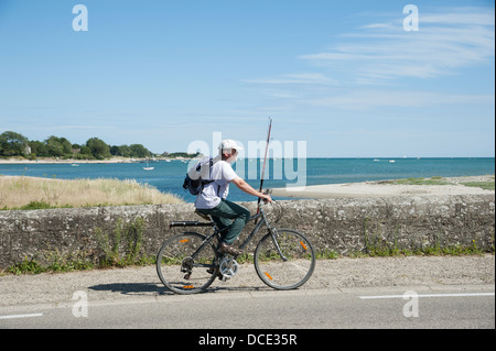 Cyclist on a bridge carrying a fishing rod over La Saire a river feeding into the sea at Reville Normandy France. Man going fish Stock Photo