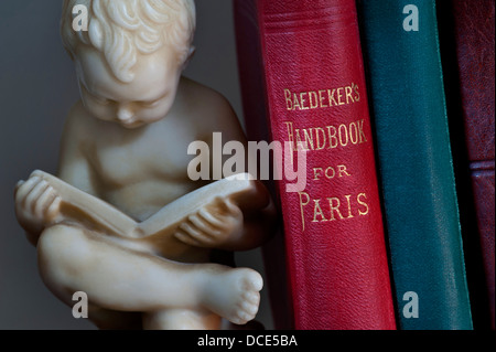 Carved bookend statue of boy reading in library situation alongside antiquarian travel books including Baedeker's guide to Paris Stock Photo