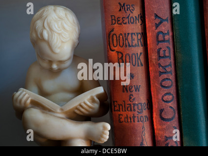 Carved bookend statue of boy reading alongside antique old cookery books in library Stock Photo