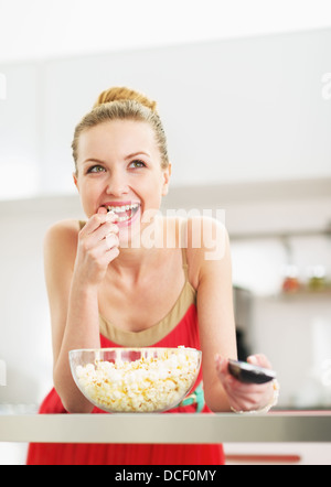 Smiling young woman eating popcorn and watching tv in kitchen Stock Photo