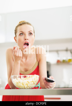 Surprised young woman eating popcorn and watching tv in kitchen Stock Photo