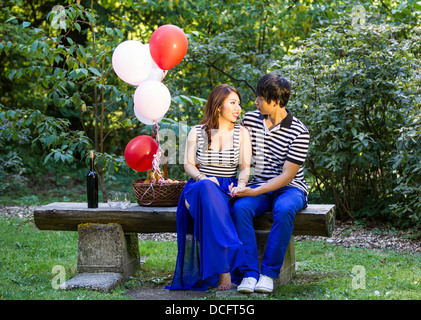 Horizontal photo of young adult couple, sitting on log bench, with balloons, basket of fruit, red wine, glasses and trees in bac