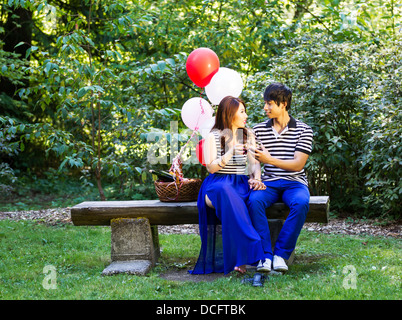 Horizontal photo of young adult couple sitting on log bench with glasses filled with red wine being held in their hands with bal