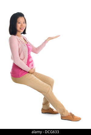 Full body six months pregnant Asian woman sitting on invisible chair, hand showing blank space looking at camera, isolated on white background. Stock Photo