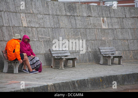 Lyme Regis, UK 17 August 2013. Tourists brave the wet weather. Couple in brightly coloured jackets sitting on bench by the Cobb Cob harbour wall. Credit:  Carolyn Jenkins/Alamy Live News Stock Photo