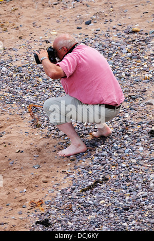 Middle aged man crouching barefoot on a beach taking photos with a camera Stock Photo