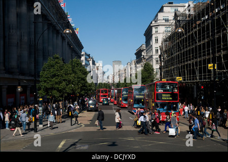 Tourist, shoppers and workers cross Oxford Street, London on a sunny day. A line of buses travel down the road. Stock Photo