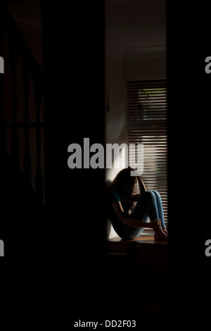 A teenage girl, hand coveing her face, sitting by a window with light pouring in. Stock Photo