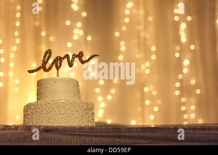 a simple white wedding cake with the word love written in sparkling gold letters on the top, & white twinkling lights background Stock Photo