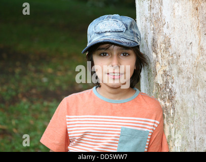 Portrait of brunette boy in baseball cap leaning against tree trunk Stock Photo