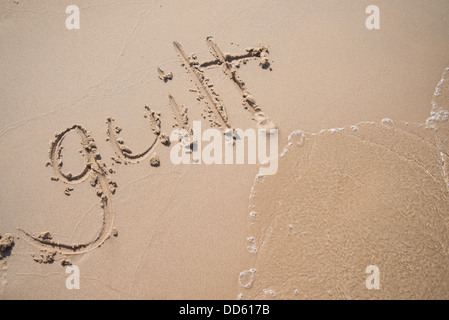 The word 'guilt' written in the sand, being washed away by a wave. Stock Photo