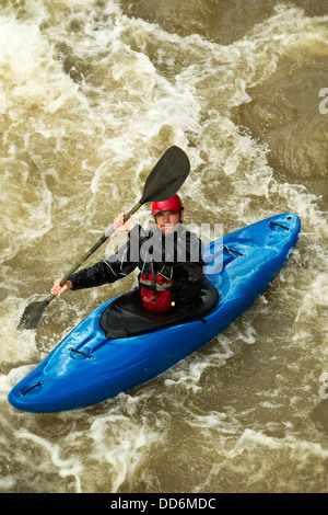 A man in a blue kayak paddles through churning, foamy whitewater rapids. He wears a red helmet, black waterproof jacket, and a red life vest, holding a black paddle mid-stroke. The river water is Stock Photo