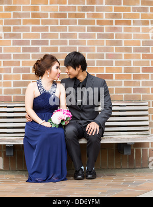 Vertical photo of young adult couple sitting on bench while looking at each other with brick wall in background Stock Photo