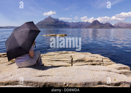 Holidaymaker under sunshade admiring view of Loch Scavaig and the Black Cuillin mountains, Elgol, Isle of Skye, Scotland, UK Stock Photo