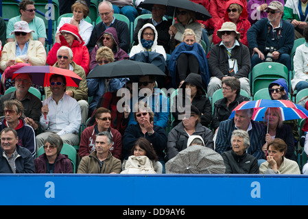 A crowd of spectators at a tennis match shelter in rain jackets and under umbrellas during a break in play Stock Photo