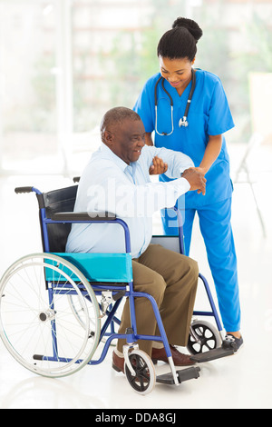 friendly African caregiver helping senior man getting up from wheelchair