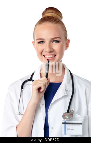 Thoughtful young female doctor or nurse looking at the camera with a lovely smile while holding her pen to her chin, upper body on white Stock Photo