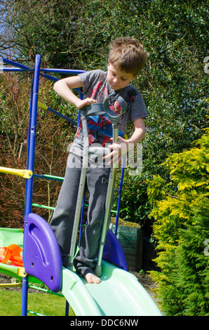 eight year old boy with crutches thinking about being adventurous on a garden slide. Stock Photo
