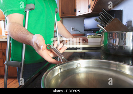 Man after anterior cruciate ligament (ACL) surgery with crutches cooking in the kitchen Stock Photo