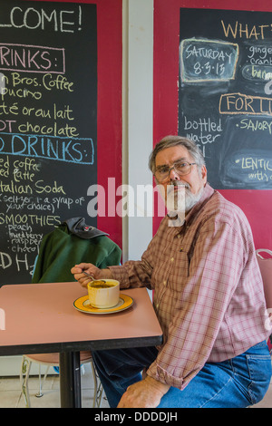 An older gray haired man sits at a restaurant table eating his soup for lunch and faces forward with spoon in his hand. Stock Photo
