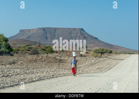 A woman with a bucket on her head standing at a gravel road in dry landscape, Kunene Region, Namibia Stock Photo