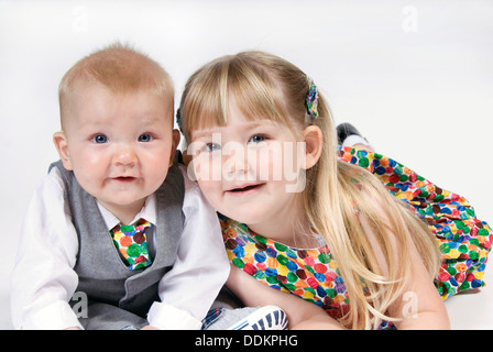Little girl lying on her tummy snuggles up to her baby brother, dressed in suit and tie and matching spotty dress, UK Stock Photo
