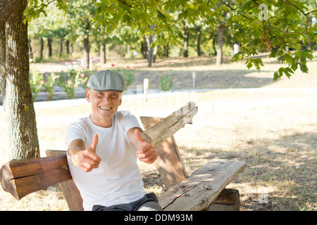 Caucasian middle-aged man showing thumbs up sitting on a bench in the park in a beautiful sunny day of summer Stock Photo