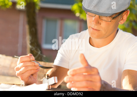 Horizontal portrait of an addicted middle-aged Caucasian man heating soluble heroin in a teaspoon with a lighter, outdoors Stock Photo