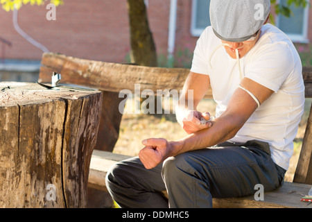 Addicted man sitting on a bench outdoors injecting his left arm intravenously Stock Photo
