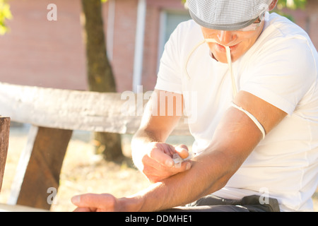 Addicted jobless middle-aged man Injecting a drug intravenously into his left arm sitting on a bench outdoors Stock Photo