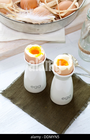 Two soft-boiled eggs in egg cups next to a bowl of fresh eggs. Stock Photo