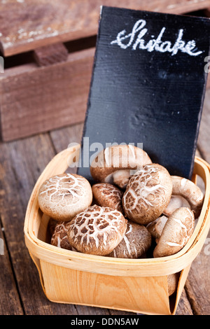 A bamboo punnet of fresh shitake mushrooms - labeled with white chalk on a small blackboard - on a rustic wood surface. Stock Photo