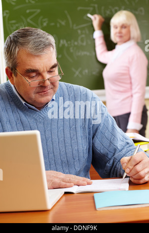 Portrait of mature man making notes on background of teacher standing by blackboard Stock Photo