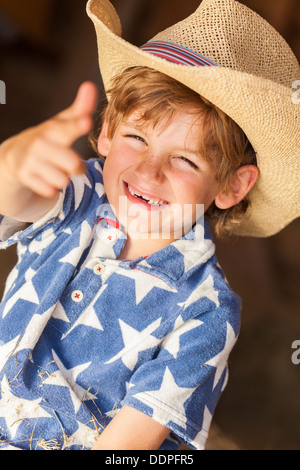 Young happy smiling blond boy child wearing a blue star shirt and cowboy hat sitting on hay or straw bales making a finger gun Stock Photo