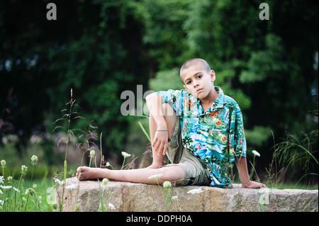 Young boy wearing a tropical style shirt sitting on a rock in a field Stock Photo