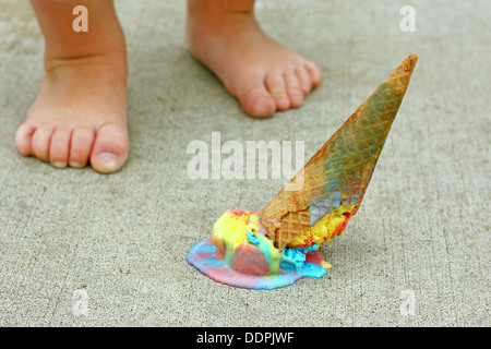 a dropped rainbow colored ice cream cone lays upside down on the sidewalk at the feet of a young child Stock Photo