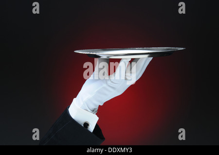 Closeup of a waiter's hand holding a silver serving tray in his fingertips over a light to dark red background. Stock Photo