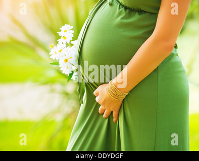 Closeup on tummy of pregnant woman, wearing long green dress, holding in hands bouquet of daisy flowers outdoors, new life Stock Photo