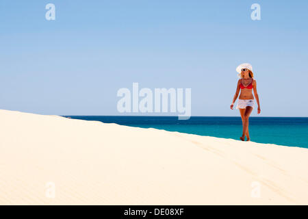 Woman wearing a hat and a bikini walking along a sandy beach Stock Photo
