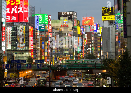 Train passing buildings lit with neon advertising signs, and traffic on a busy road, Shinjuku District, Tokyo, Japan Stock Photo