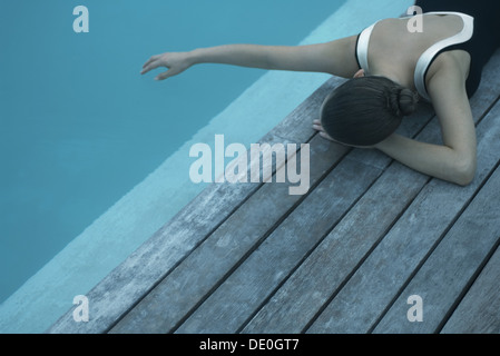 Woman in swimsuit lying on deck next to swimming pool, touching surface of water with hand Stock Photo