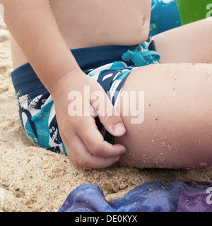 Little boy sitting on sand, scratching leg, cropped