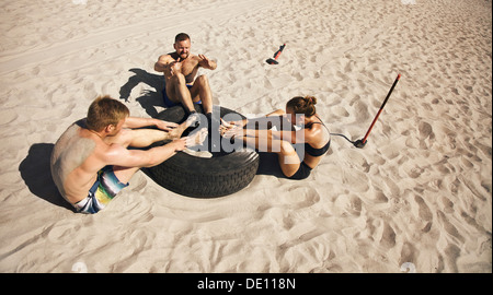 Fitness and healthy lifestyle. Small group of young athletes doing abdominal exercise with a truck tire on beach. Athletes doing Stock Photo