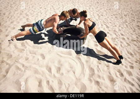 Small group of people doing push-ups on tire. Young athletes working out on beach during a hot summer day. Stock Photo