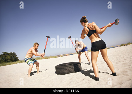 Three strong athletes doing hammer strike on a truck tire during crossfit exercise outside on beach Stock Photo